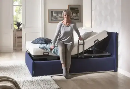 Seniorin sitzt auf modernem Pflegebett mit Aufstehhilfe im hellen Schlafzimmer.