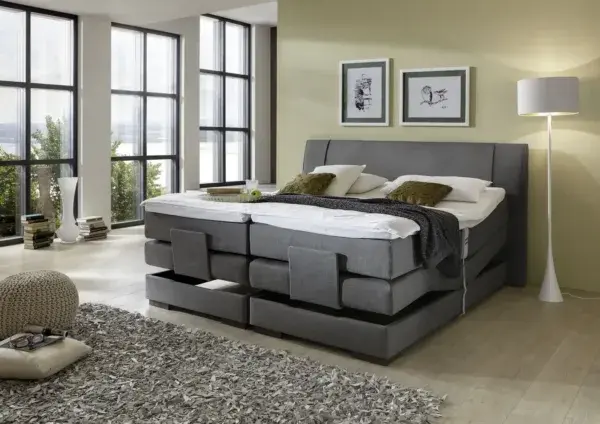 Modernes Boxspringbett im hellen Schlafzimmer mit großen Fenstern und stilvoller Einrichtung.