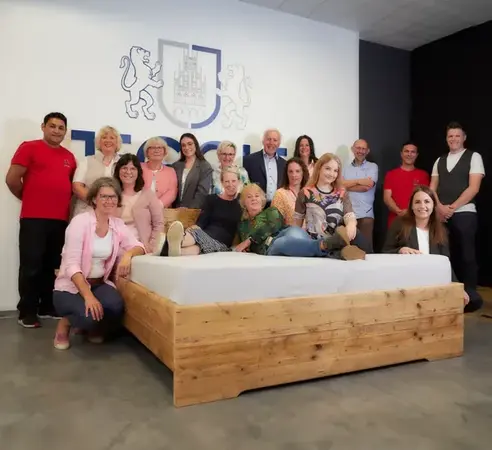 Gruppe von Menschen posiert in einem Ausstellungsraum um ein Massivholzbett mit Lattenrost.