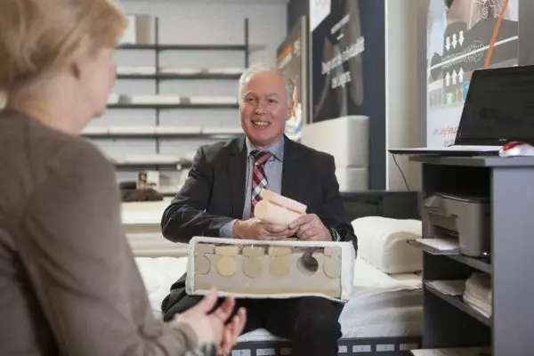 Berater erklärt Kundin im Matratzenfachgeschäft den Aufbau einer Matratze am Modell.