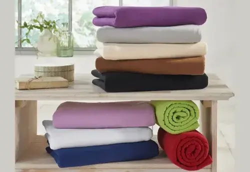 Mehrere bunte Fleecedecken liegen ordentlich gefaltet auf einem Holzregal im Wohnzimmer.