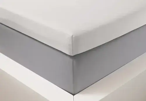 Ecke eines Bettes mit grauem Spannbettlaken und weißer Matratze, modernes Schlafzimmer.